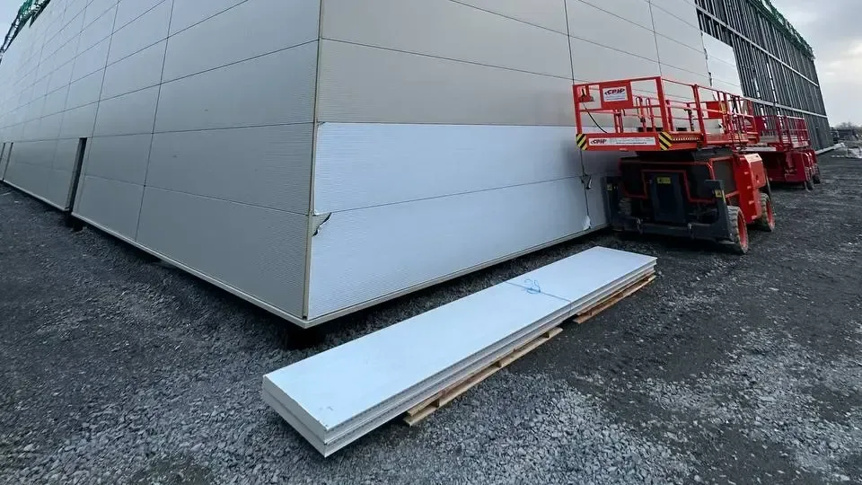 Insulated wall panels on a pallet in front of a freshly clad industrial building corner, with a mobile elevating work platform