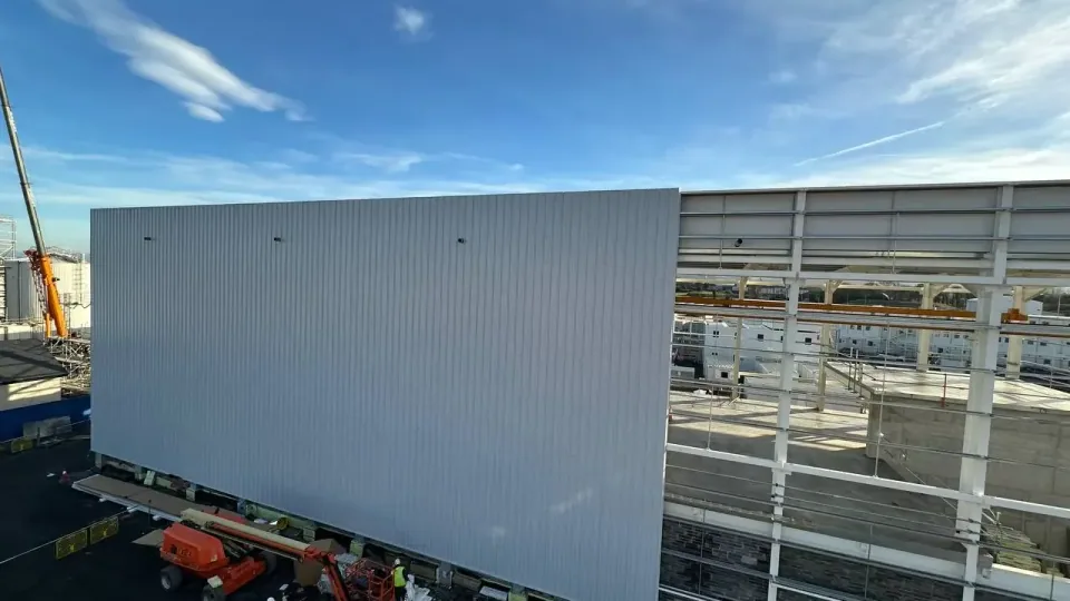 Grey insulated wall panels installed on one elevation of an industrial building, the next bay still open steel frame, a work platform below